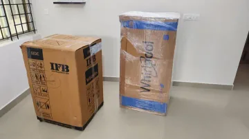 A washing-machine carton beside another tall appliance package with outer wrapping.