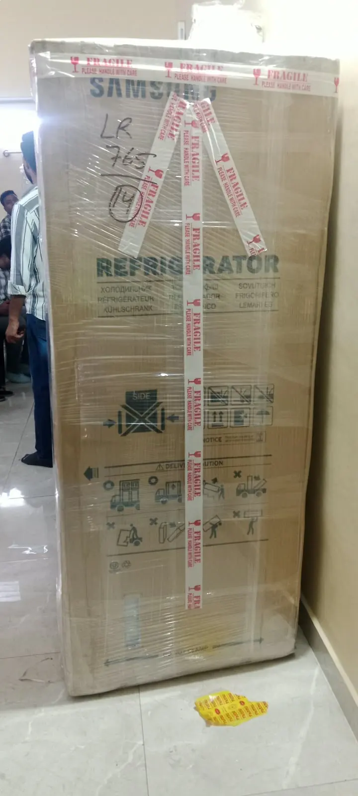 A tall household item enclosed in cardboard protection and outer wrapping.