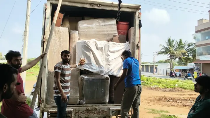 People beside the open rear of a goods vehicle containing wrapped furniture and household packages.