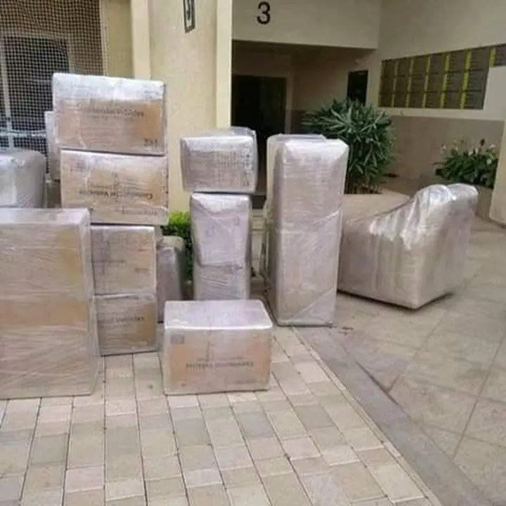 Wrapped cartons and furniture pieces grouped on paving outside a building.